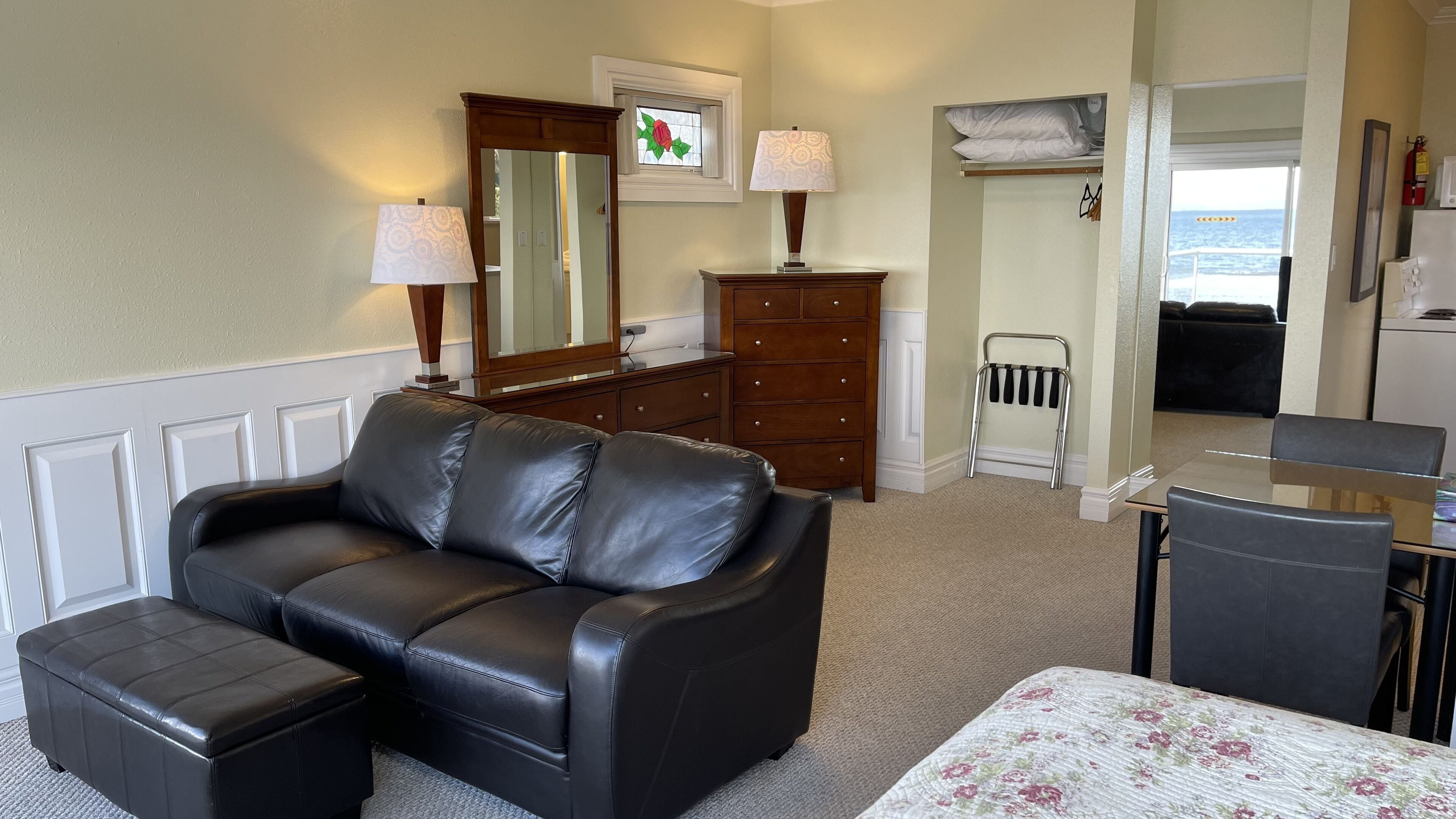 Habitación de hotel con sofá de cuero negro, un pequeño reposapiés, cómoda de madera, espejo, ventana con adorno de rosas, mesita de noche con lámpara, cama con colcha estampada, pequeña mesa con sillas y extintor.