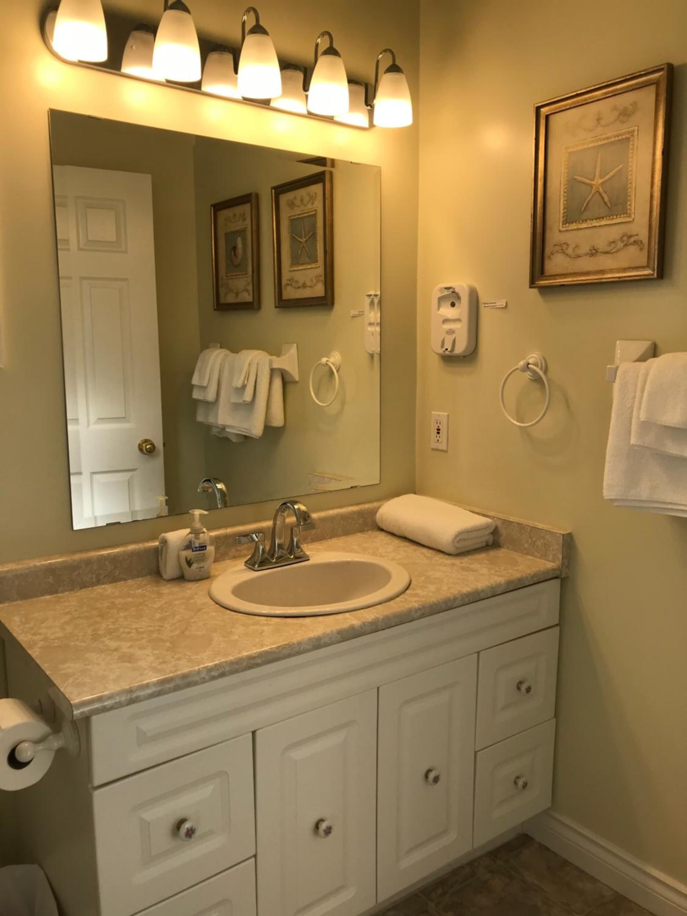 A bathroom with a large mirror, a sink, and a vanity with drawers. There are towels hanging on the wall and a framed picture above the mirror.