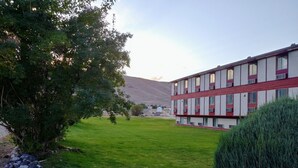 A building with a red and white facade, a grassy courtyard, and a mountain in the background. - Northgate Inn Motel (Challis)