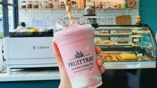 A hand holding a pink smoothie with a straw, labeled 'FRUITTRIO', in a cafe with a white brick wall and a sign above a counter displaying various food items. - Phaithong Sotel Resort (Chalong)