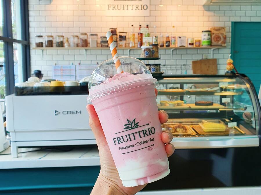 Una mano sostiene un batido rosa con una pajita, etiquetado 'FRUITTRIO', en una cafetería con una pared de ladrillo blanco y un cartel sobre un mostrador que exhibe diversos alimentos.