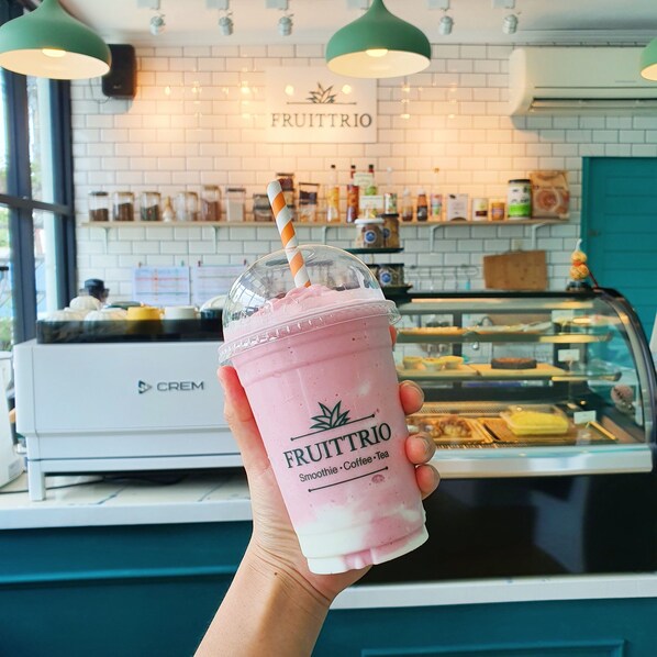 Mão segurando um smoothie rosa com canudo, identificado como 'FRUITTRIO', em um café com parede de tijolos brancos e um letreiro sobre o balcão exibindo diversos itens de comida.