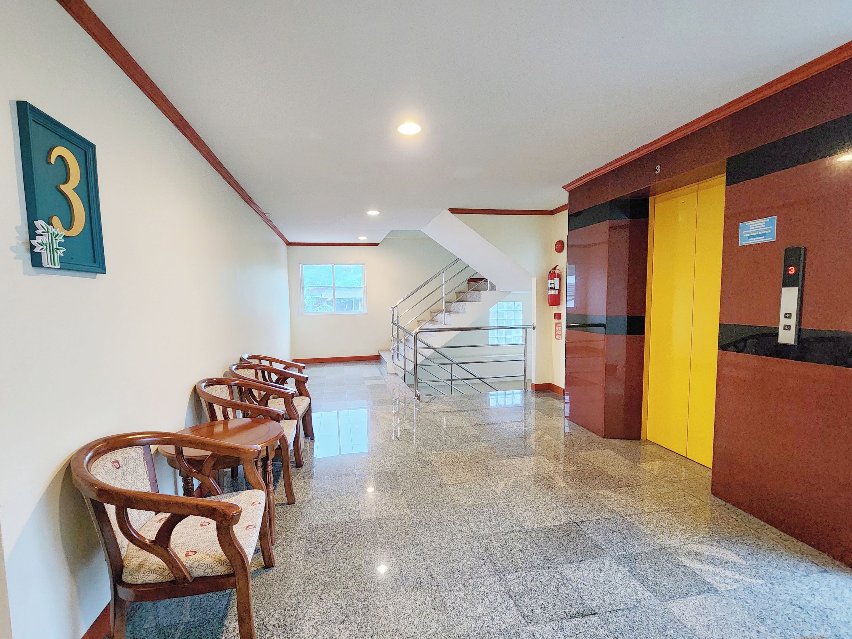 Un hall d’hôtel avec un escalier, des chaises en bois, un extincteur et un panneau indiquant le numéro de la chambre, fixé au mur.