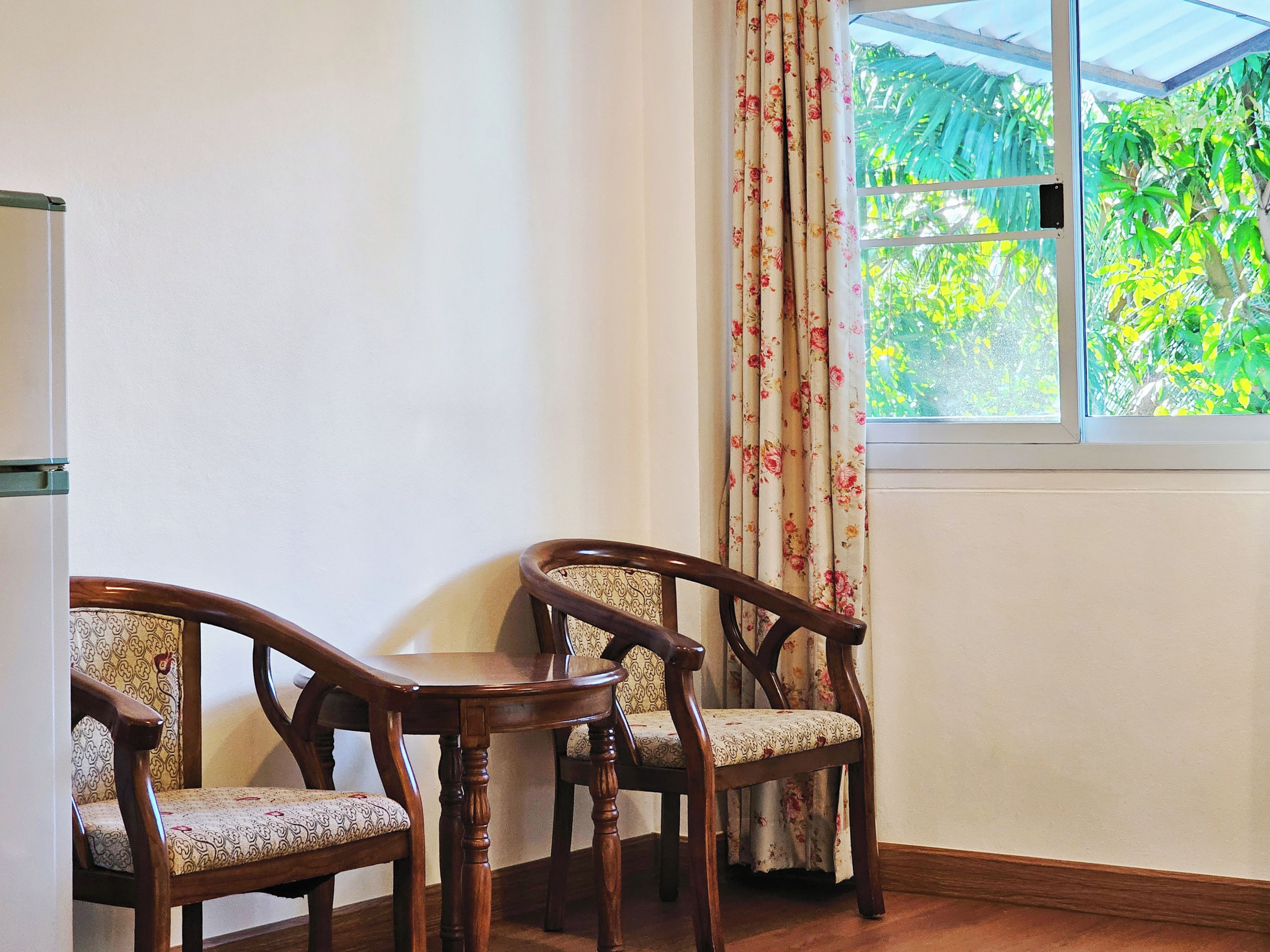 A room with two wooden chairs and a small table, a window with curtains, and a white wall.