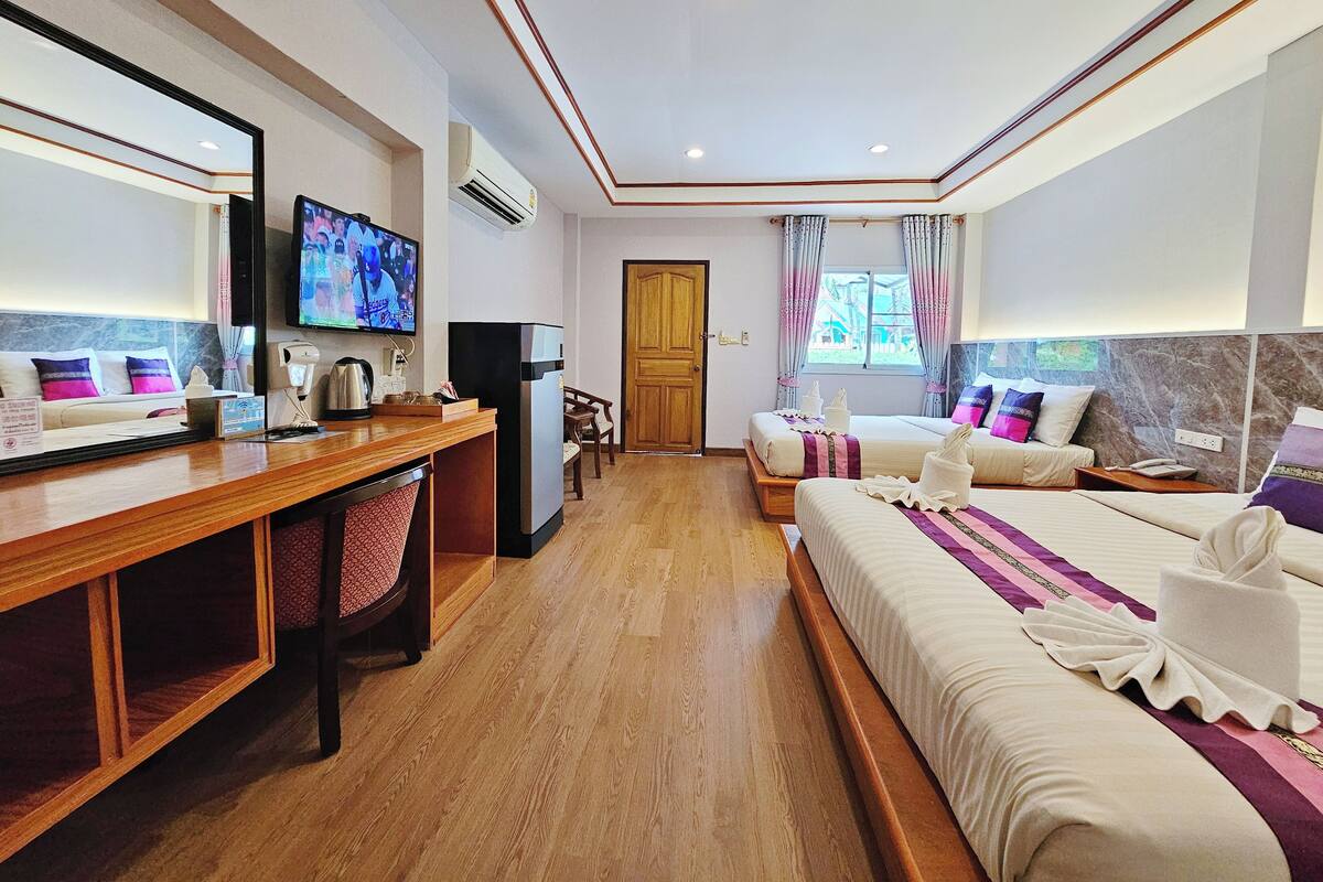 Habitación de hotel con escritorio de madera, televisor, cama con cubrecama morado y blanco, y ventana con cortinas.