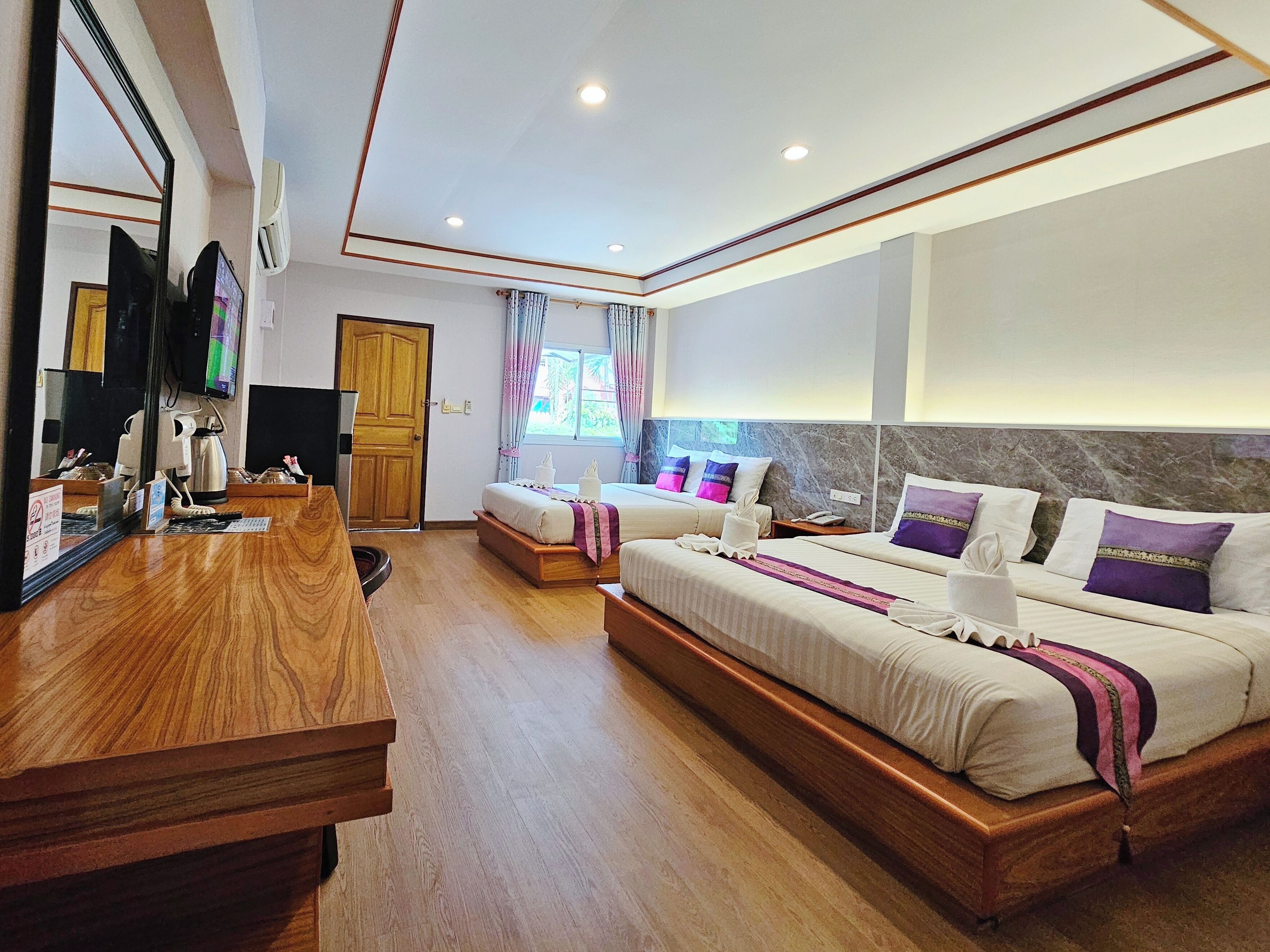 Habitación de hotel con un escritorio de madera, dos camas con almohadas moradas, un espejo grande y un televisor.