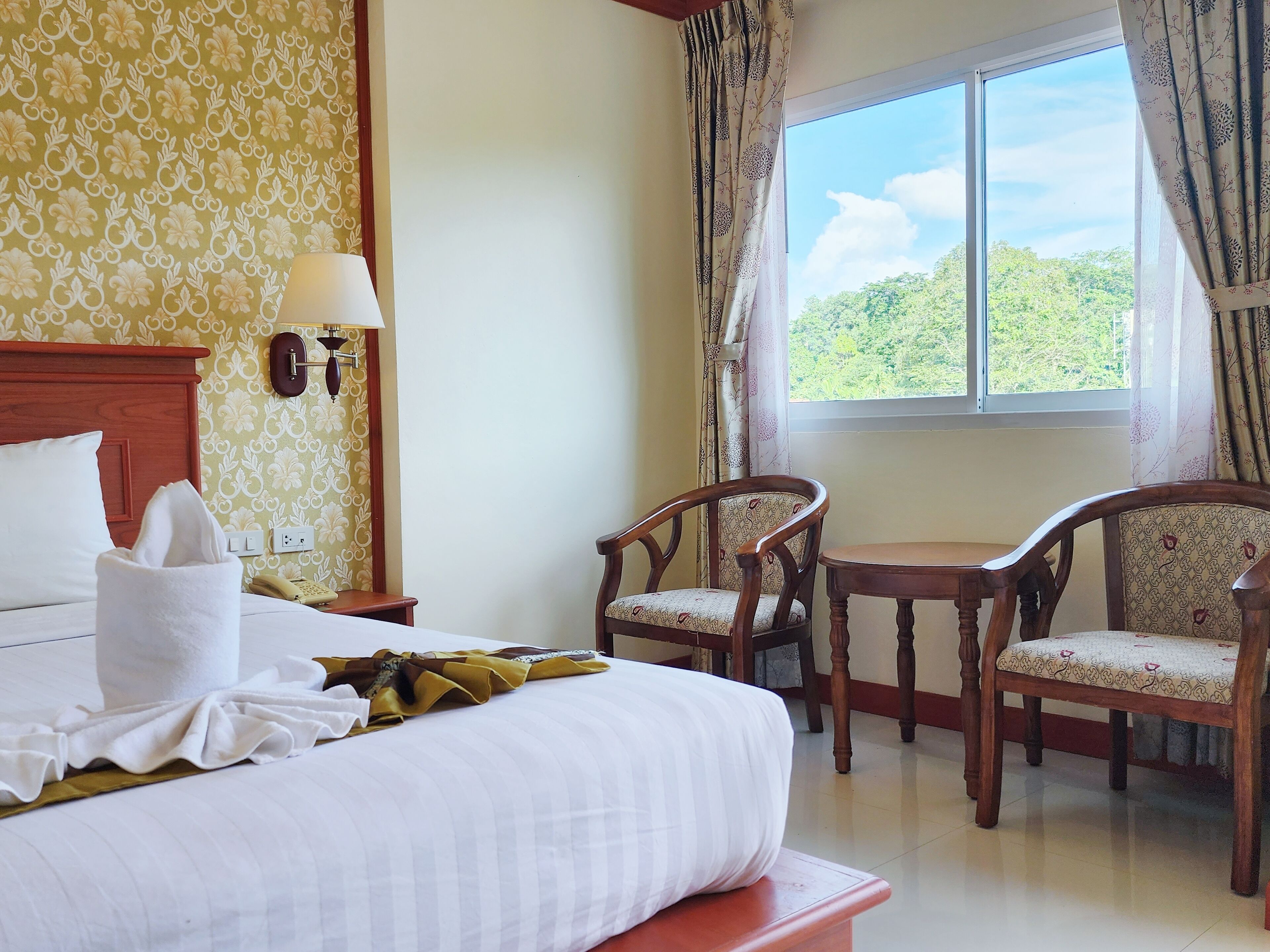 A hotel room with a bed, two chairs, a table, a lamp, and a window with a view of greenery.
