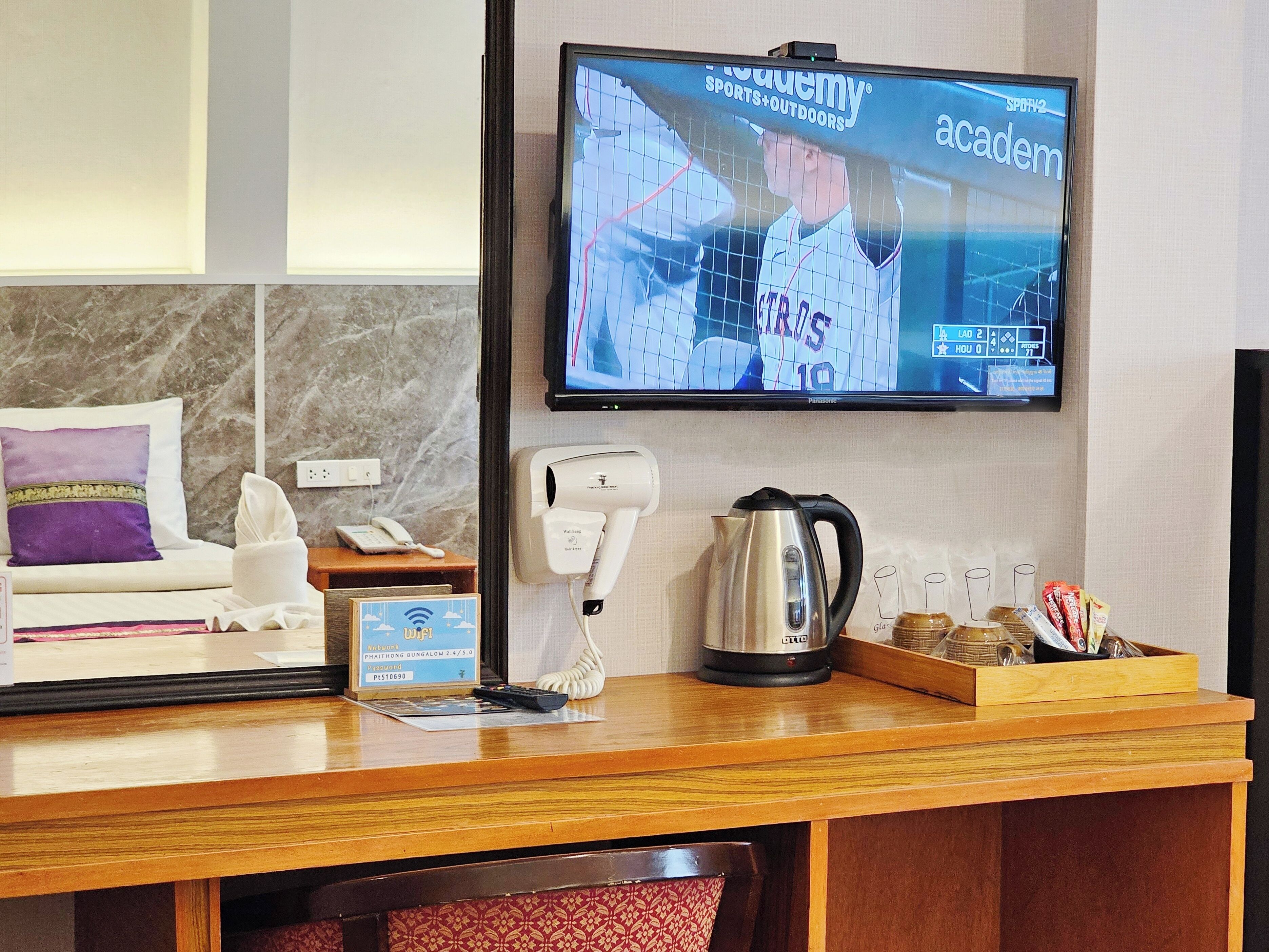 Zona de recepción de un hotel con un televisor de pantalla plana mostrando una transmisión deportiva, un teléfono, un cartel de Wi-Fi, un hervidor de agua y una bandeja con aperitivos.