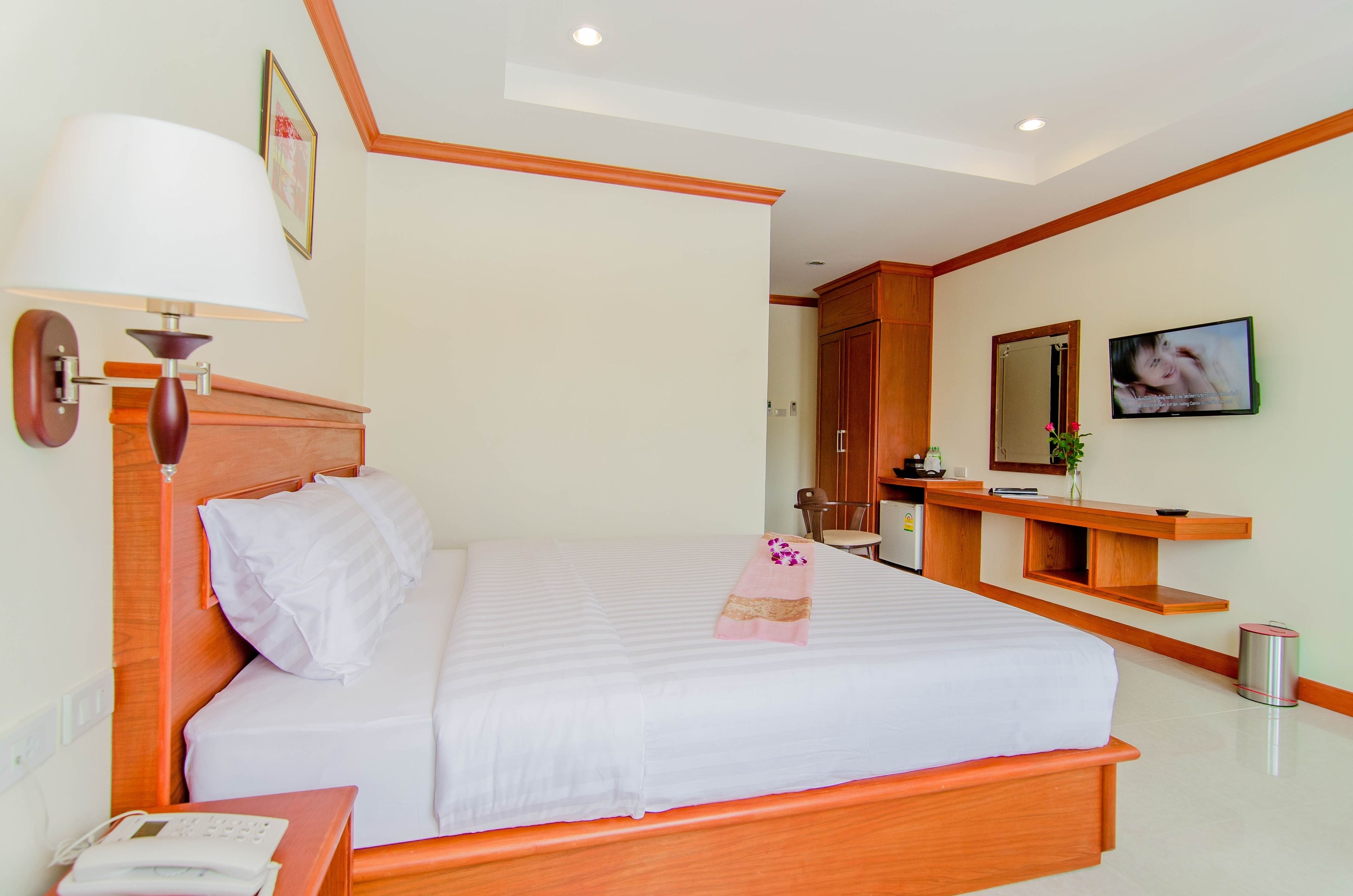 A neatly made bed with a white comforter and pillows, a wooden headboard, a bedside lamp, a small table with a phone, and a television on the wall.