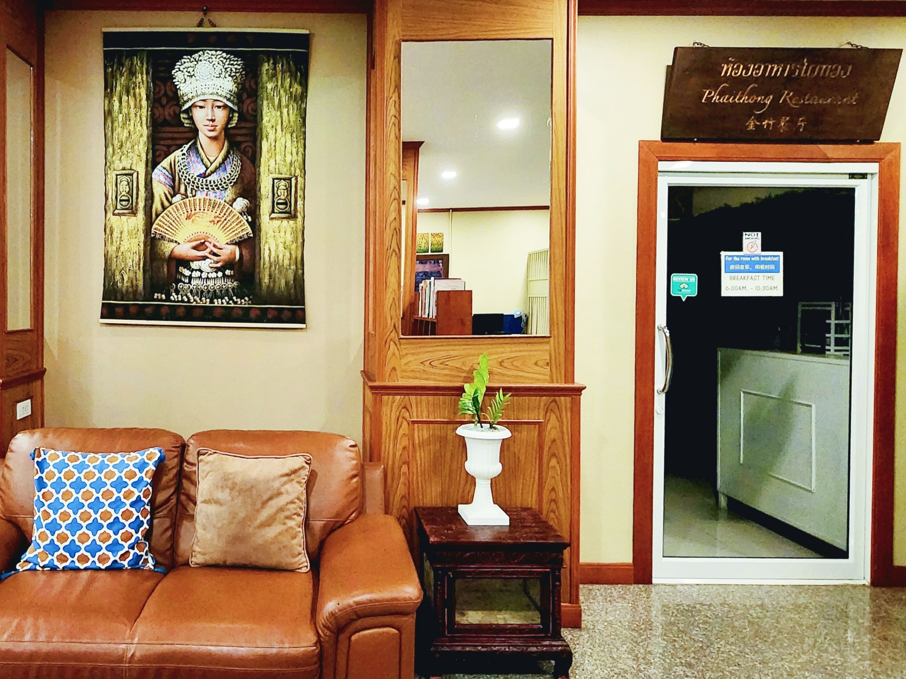 A lobby with a brown leather sofa, a wooden cabinet with a mirror, a framed picture on the wall, and a sign indicating breakfast hours.