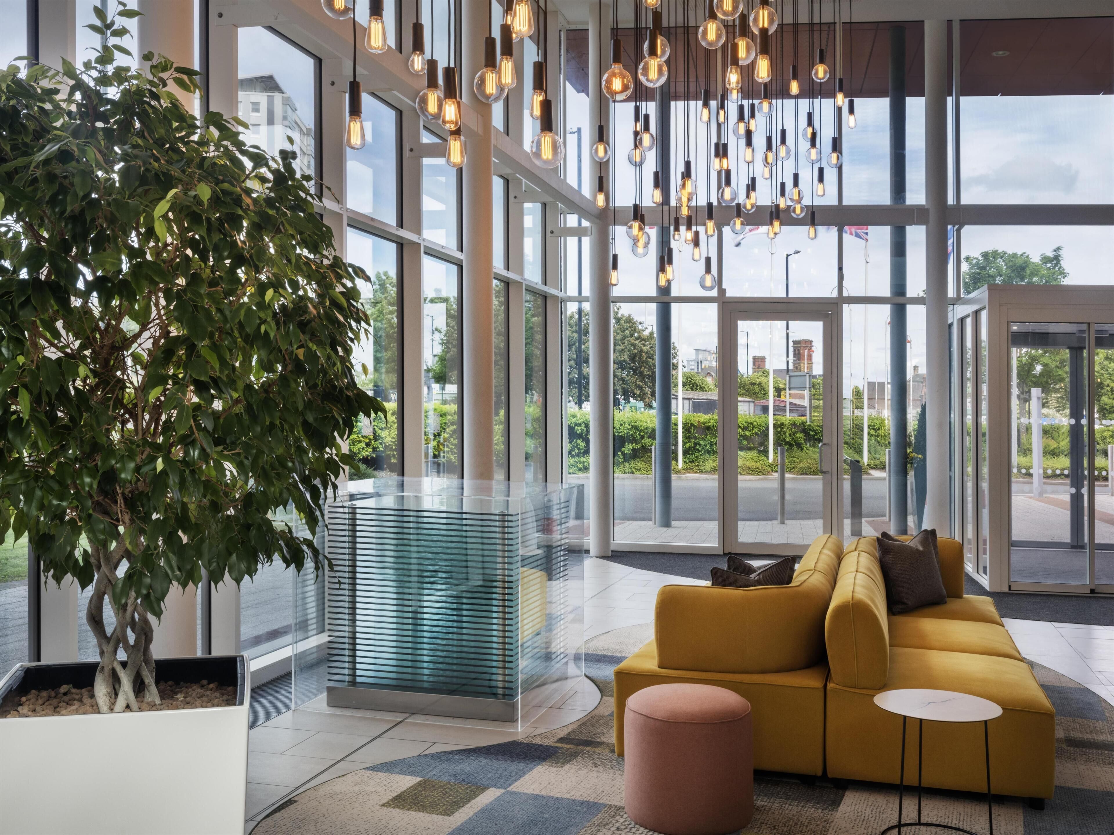A modern lobby with a large glass partition, a yellow sofa, a round ottoman, and hanging light fixtures.