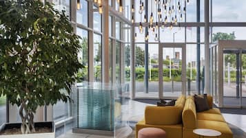 A modern lobby with a large glass partition, a yellow sofa, a round ottoman, and hanging light fixtures.
