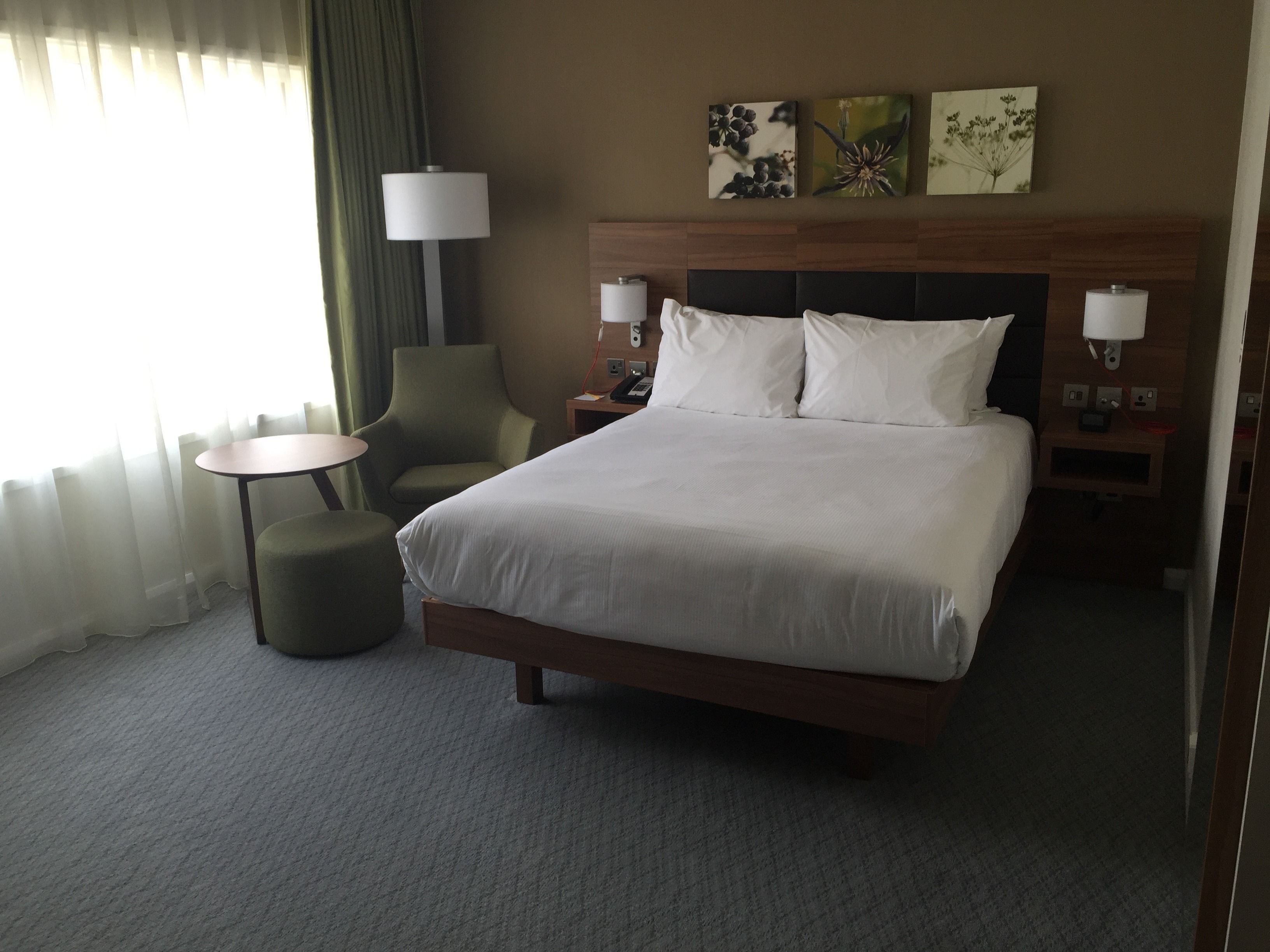 Una camera d'albergo con un letto grande, due comodini con lampade, una sedia, un tavolino rotondo e una finestra con tende leggere.