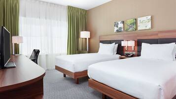 A hotel room with two beds, a desk, a chair, a TV, and a lamp.
