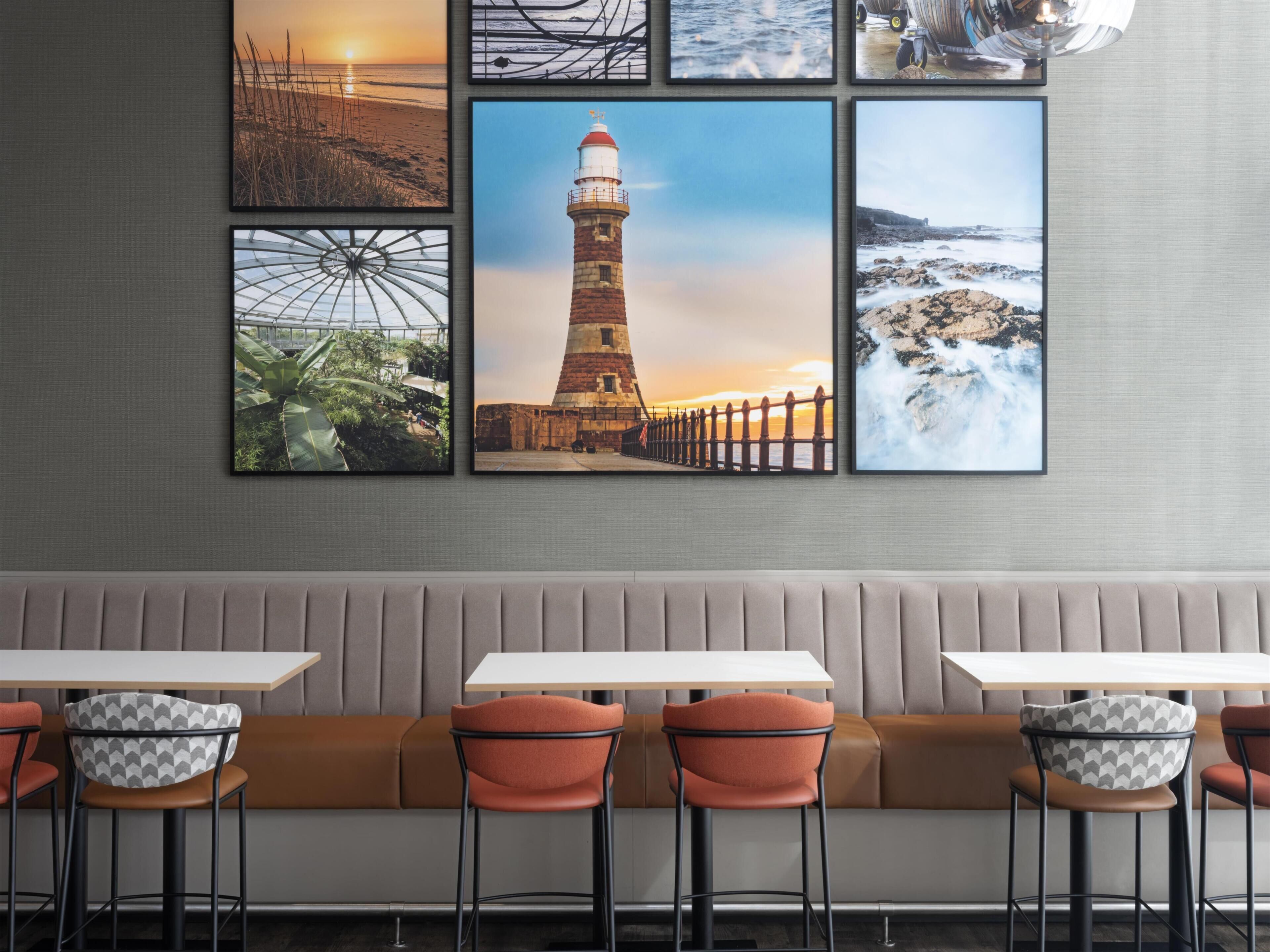 A restaurant interior with a long bench seating area, multiple tables, and a wall adorned with framed coastal landscape photographs.