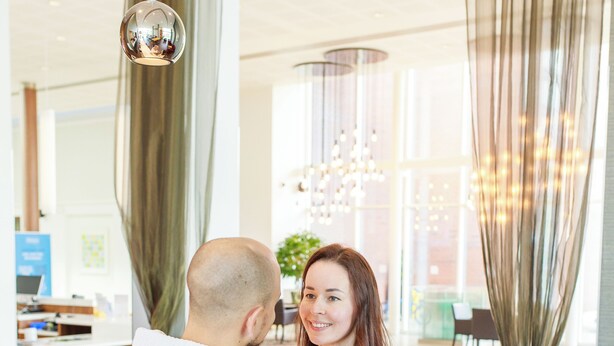 A modern interior with a wooden dining table, a hanging spherical light fixture, and a large window with curtains. - Hilton Garden Inn Sunderland (Sunderland)