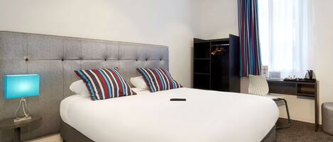 Habitación de hotel con una cama grande, una mesita de noche con lámpara, un armario y una ventana con cortinas.