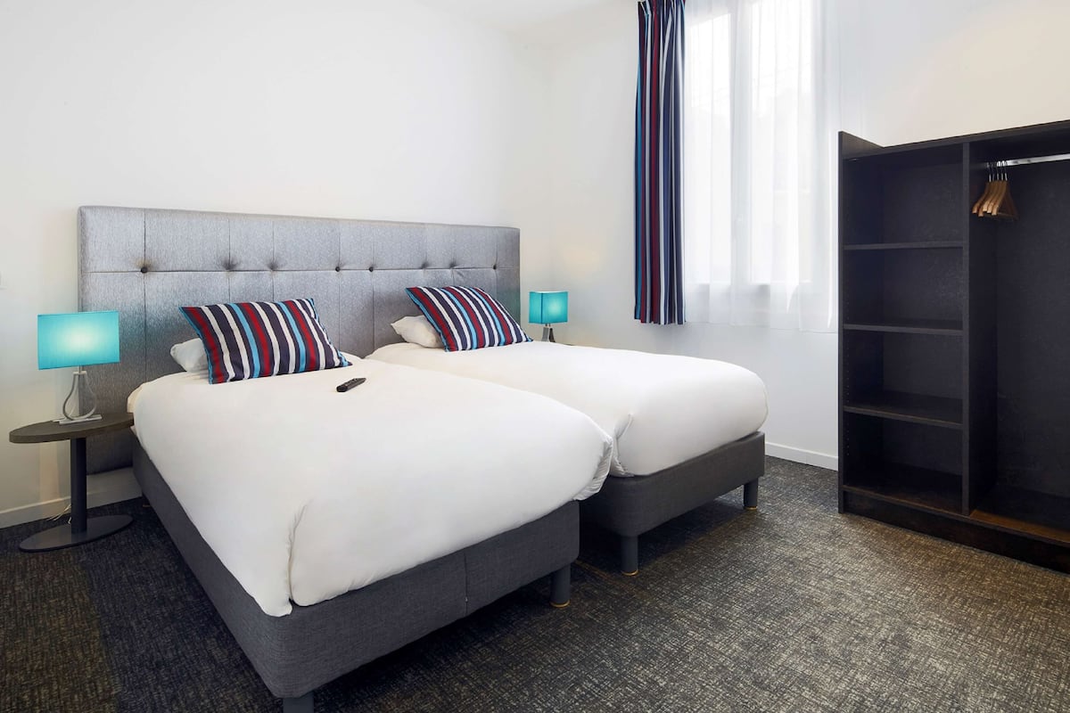 A hotel room with two beds, a grey headboard, striped pillows, a blue lamp, a wardrobe, and a window with sheer curtains.