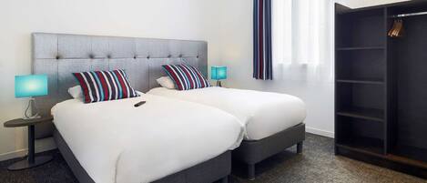 Une chambre d’hôtel avec deux lits, une tête de lit grise, des oreillers à rayures, une lampe bleue, une armoire et une fenêtre avec des rideaux légers.
