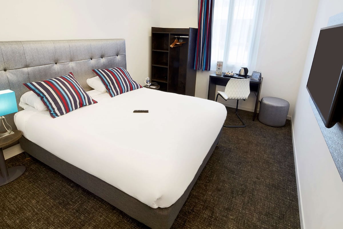 A hotel room with a large bed, a desk, a chair, a TV, and a small stool.