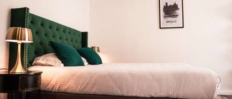 Uma cama bem arrumada com cabeceira verde, uma luminária de cabeceira e um quadro com uma vista urbana na parede.