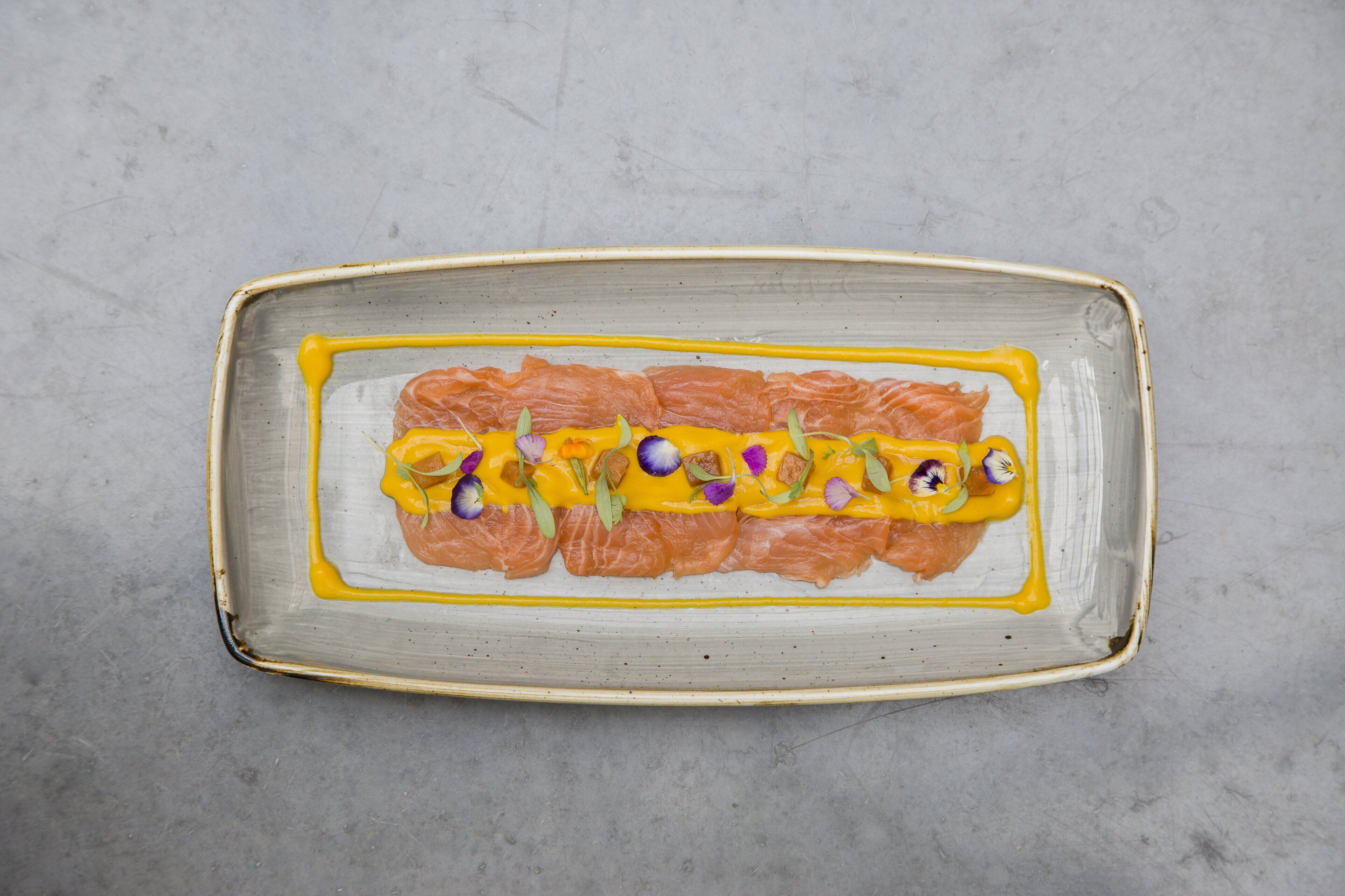 A rectangular ceramic plate with salmon and yellow sauce, garnished with purple flowers.