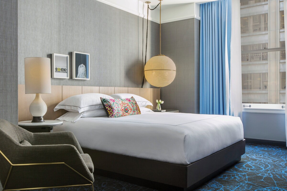 Une chambre d’hôtel avec un grand lit, une table de chevet avec une lampe, une chaise et une vue sur un immeuble par la fenêtre.
