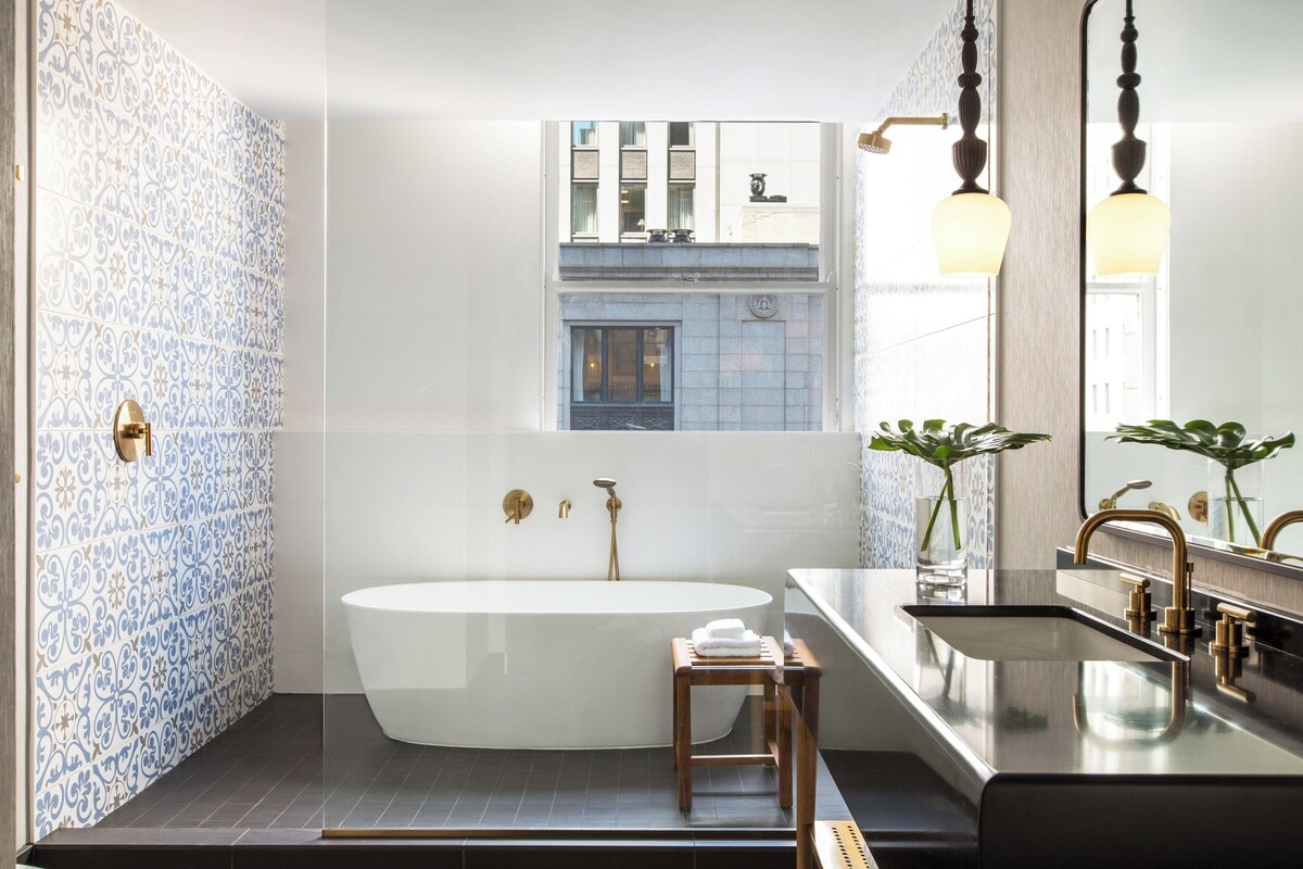 Une salle de bain moderne avec une baignoire sur pieds, un meuble-lavabo avec un miroir et une vue sur un immeuble par la fenêtre.