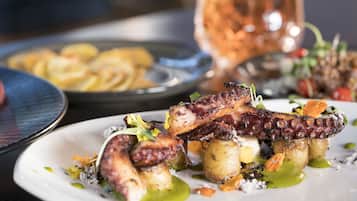 A plate of octopus with potatoes and greens, a glass of wine, and a metal dish in the background.