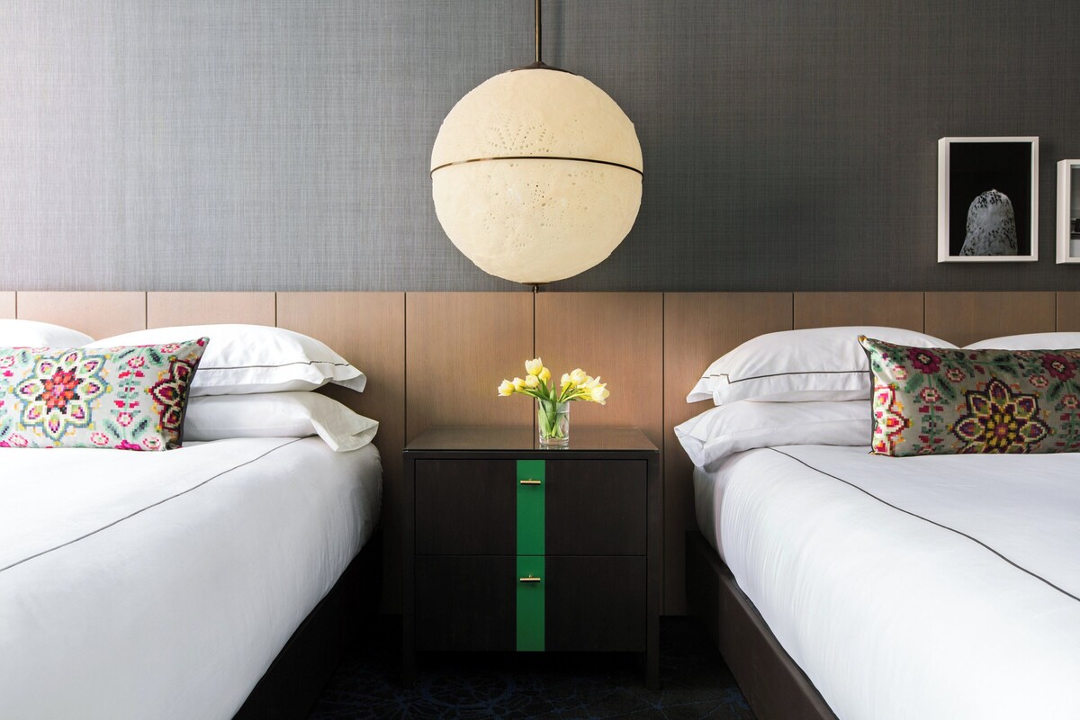 Une chambre d’hôtel avec deux lits, une commode verte, un luminaire suspendu et un vase avec des fleurs sur la table de chevet.
