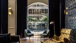 Un salon moderne comprenant un canapé, deux fauteuils, une chaise longue, une table ronde et une vue sur un immeuble par une grande fenêtre.