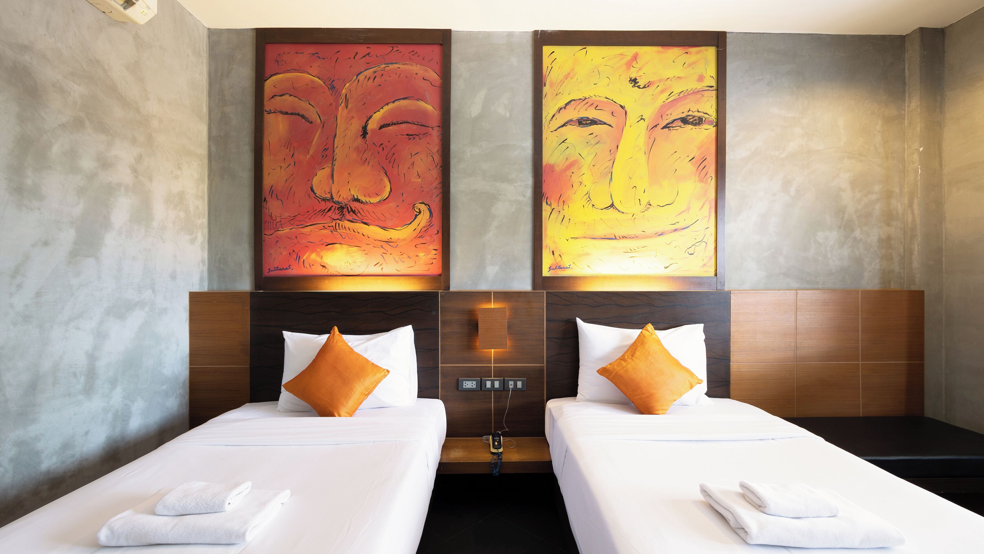 A hotel room with two beds, each with white linens and orange pillows. There are abstract paintings on the wall above the beds.