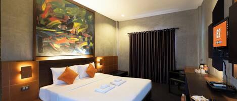 Habitación de hotel con un cuadro abstracto grande, una cama con sábanas blancas y almohadas naranjas, un escritorio con una computadora portátil y un televisor montado en la pared.