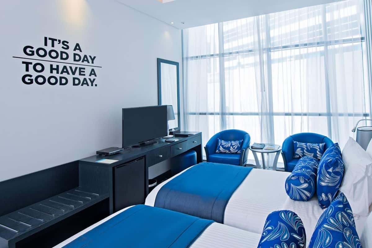 A modern hotel room with a large bed, two armchairs, a desk, and a TV. There's a large window with sheer curtains and a motivational quote on the wall.
