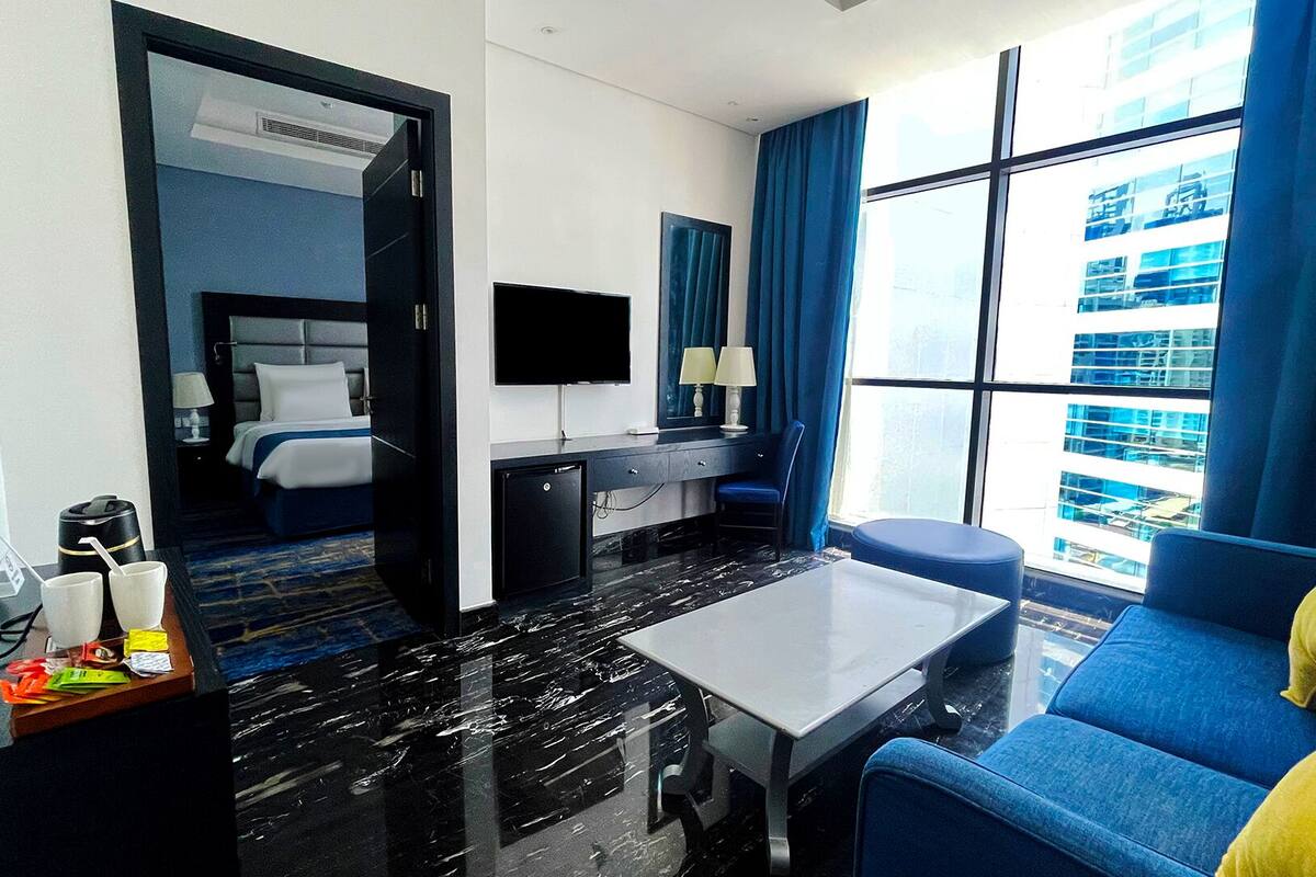 A hotel room with a bed, desk, TV, and a view of the city through large windows.