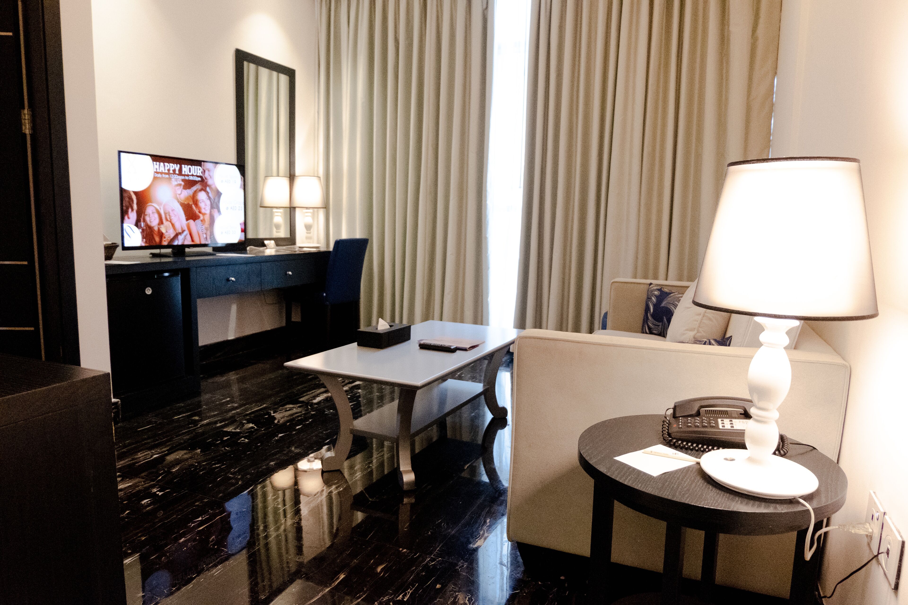 A hotel room with a flat-screen TV, a desk with a chair, a sofa, a lamp, and a telephone.