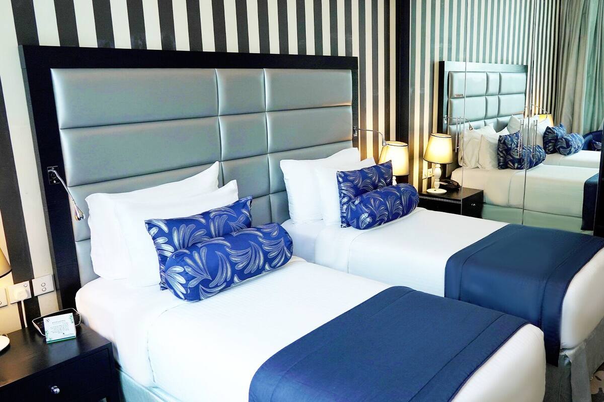 A hotel room with two beds, a large mirror, a nightstand with a lamp, and a small cabinet.