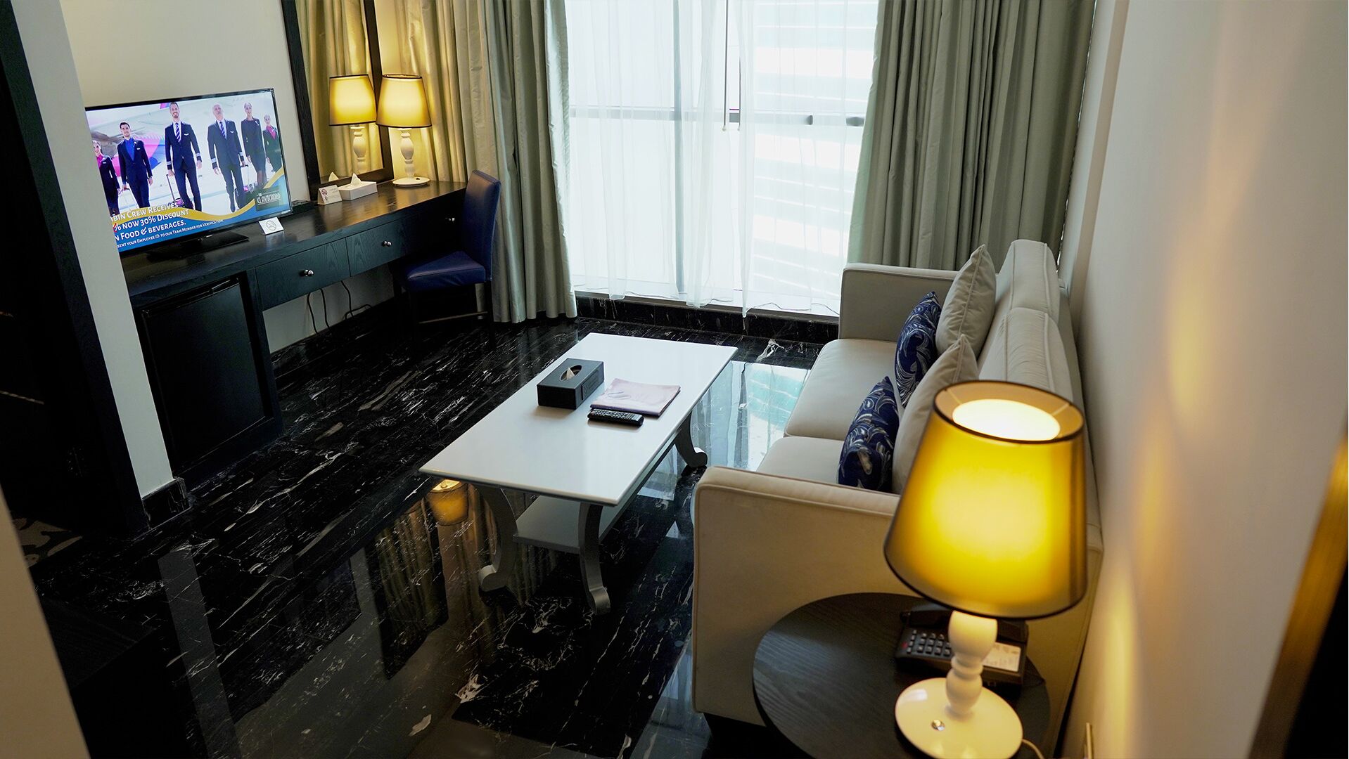 A hotel room with a TV, a sofa, a table, and a lamp.