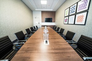 Une salle de conférence avec une longue table en bois, des chaises noires, un téléviseur à écran plat fixé au mur et des œuvres d’art encadrées accrochées au mur.