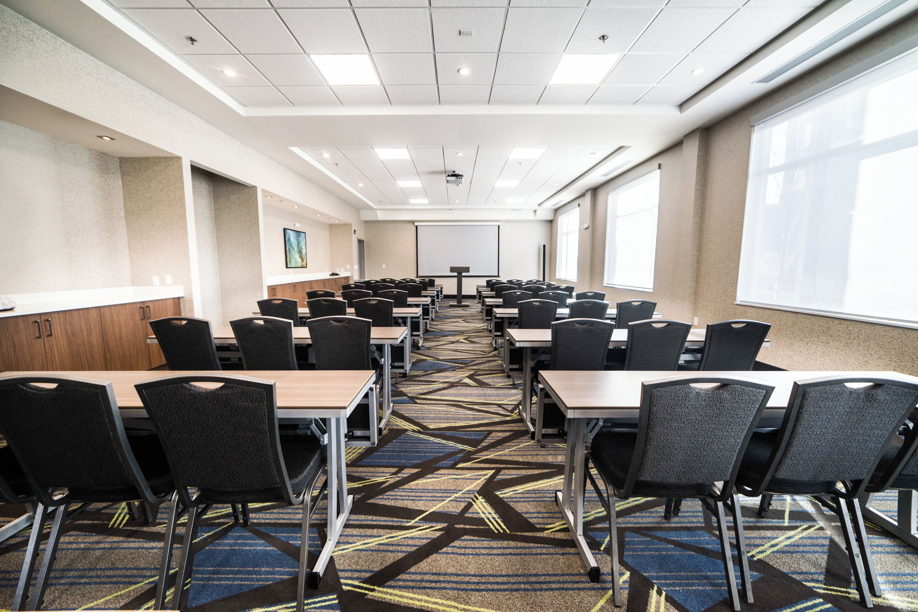 Un salón de conferencias con filas de mesas y sillas, una pantalla de proyector y ventanas amplias.