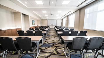 Un salón de conferencias con filas de mesas y sillas, una pantalla de proyector y ventanas amplias.