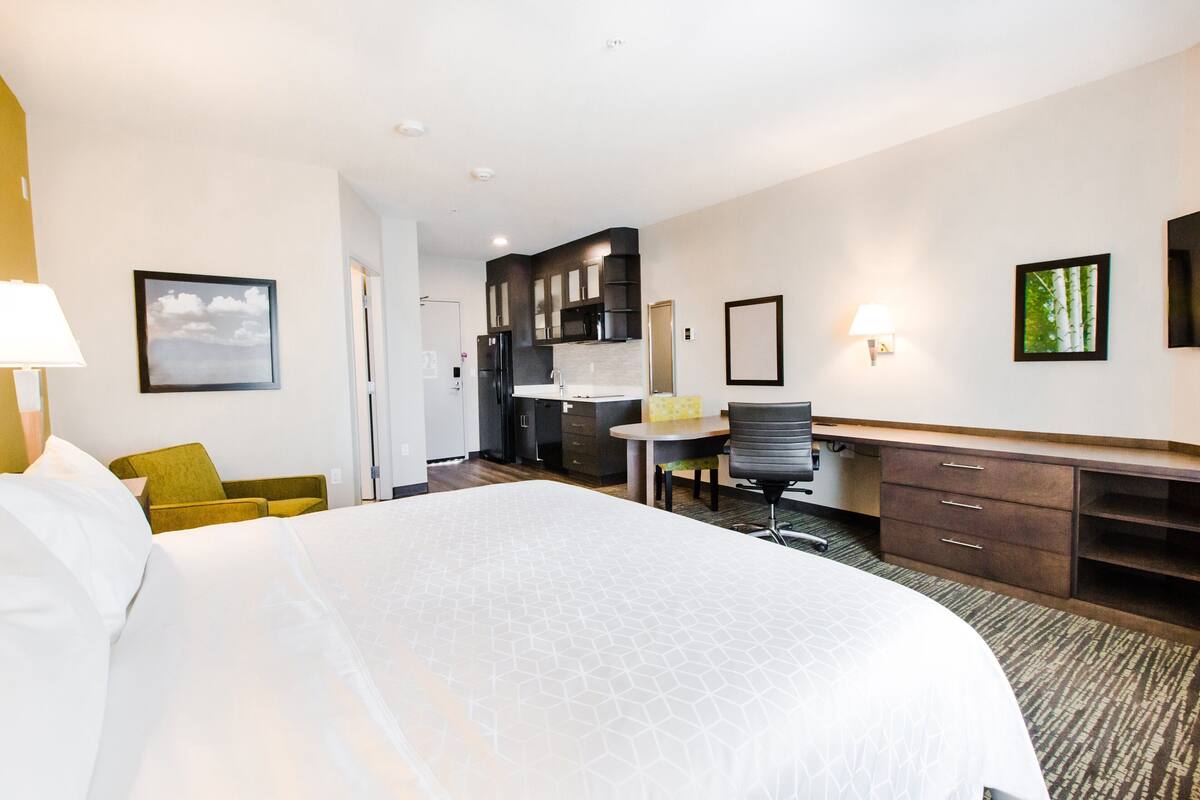Habitación de hotel con una cama grande, un escritorio, una silla, una mesita y una kitchenette al fondo.
