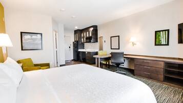 A hotel room with a large bed, a desk, a chair, a small table, and a kitchenette in the background.