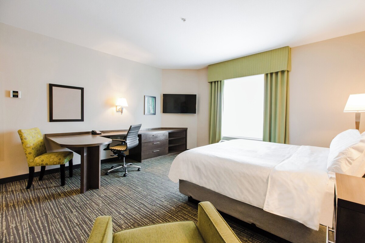 Quarto de hotel com cama, escrivaninha, cadeira, televisão e janela com cortinas.