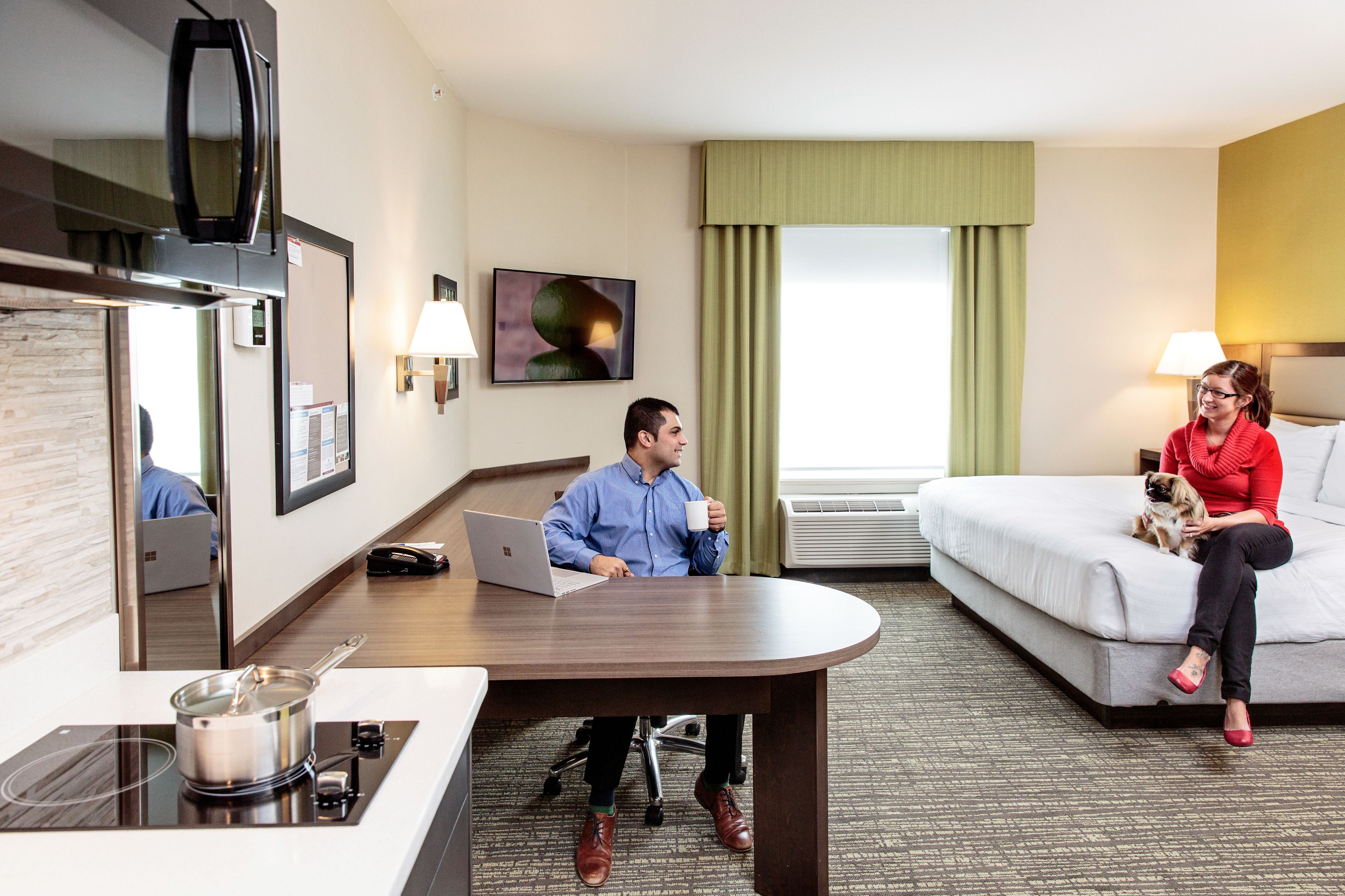 A hotel room with a desk, a bed, a sofa, a microwave, a lamp, and a painting on the wall.