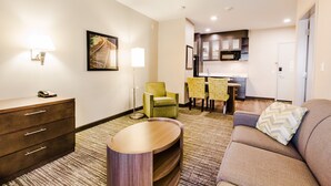 Habitación de hotel con sofá, mesita de madera, lámpara, silla, mesa de comedor con sillas y zona de cocina.