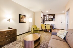 Quarto de hotel com sofá, mesa de cabeceira de madeira, luminária, cadeira, mesa de jantar com cadeiras e área de cozinha.