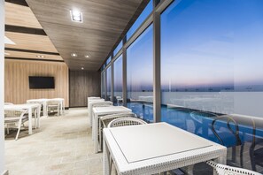 Een modern binnenzwembad met witte rieten meubels en een groot raam met uitzicht op zee.