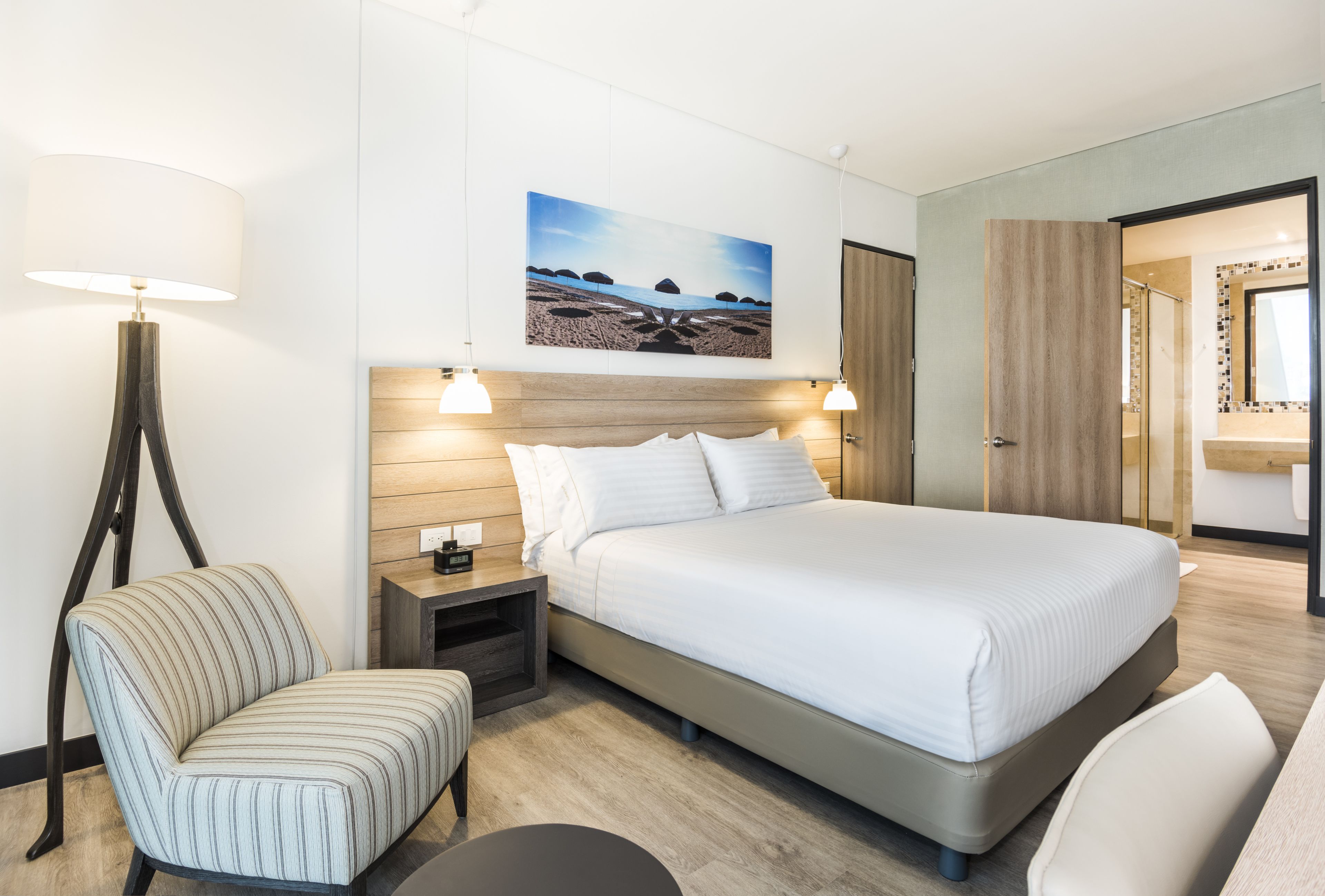 Una habitación de hotel moderna con una cama grande, una mesita de noche, una lámpara, una silla y un cuadro en la pared.