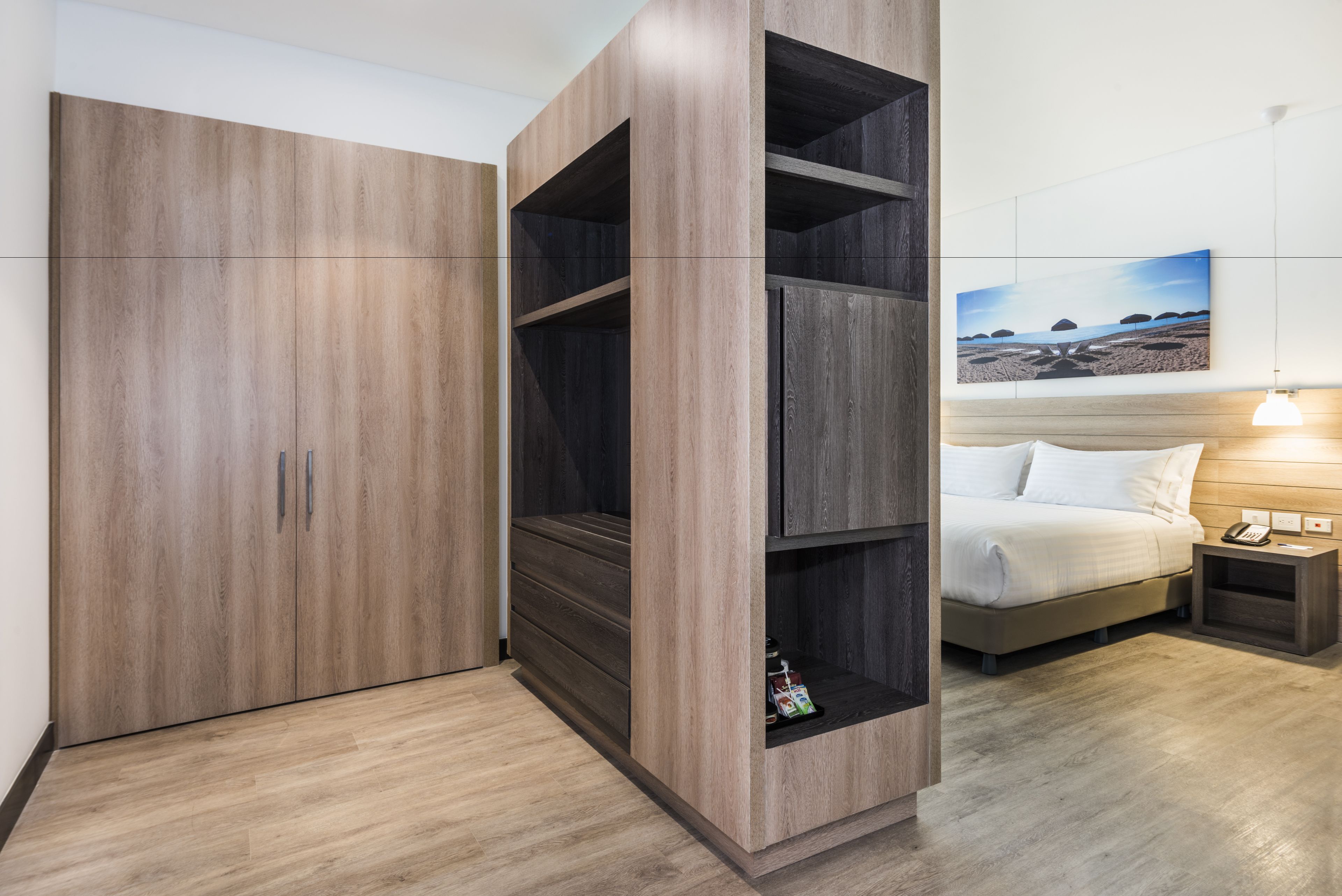 A hotel room with a large wardrobe, a bed with a headboard, a bedside table with a lamp, and a wall-mounted painting.
